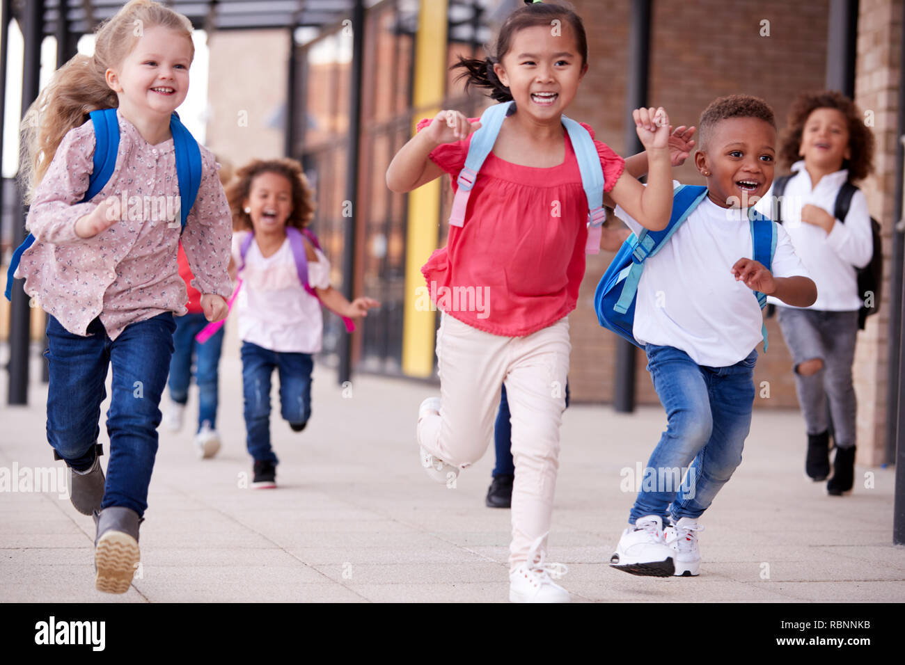 Un gruppo di sorridere multi-etnico i bambini della scuola in esecuzione in una passerella al di fuori delle loro scuole materne edificio dopo una lezione, chiudere fino Foto Stock