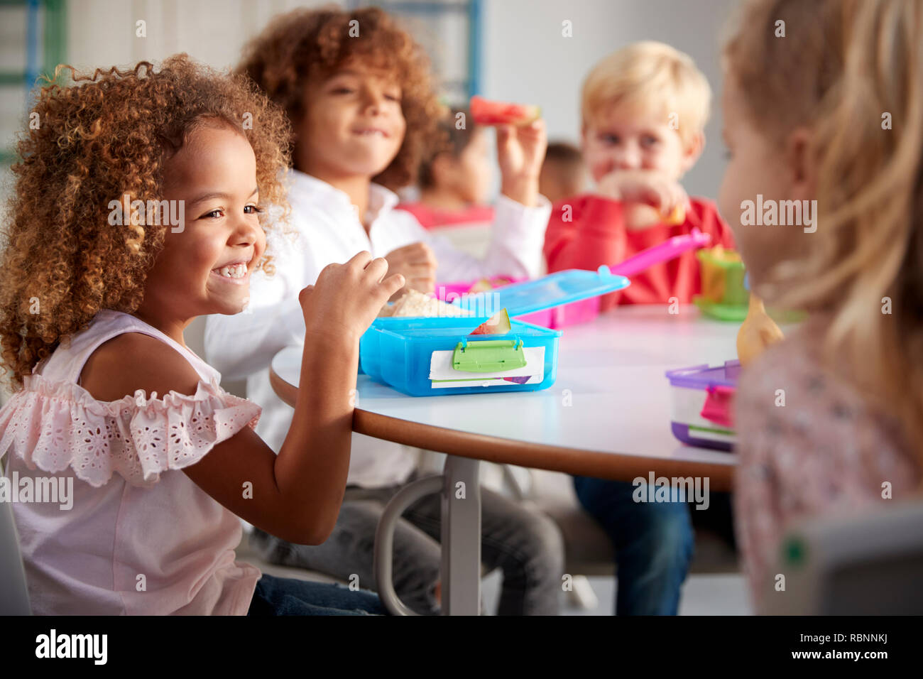 Close up di sorridere i giovani bambini seduti ad un tavolo a mangiare il loro pranzo al sacco insieme alla scuola infantile, il fuoco selettivo Foto Stock