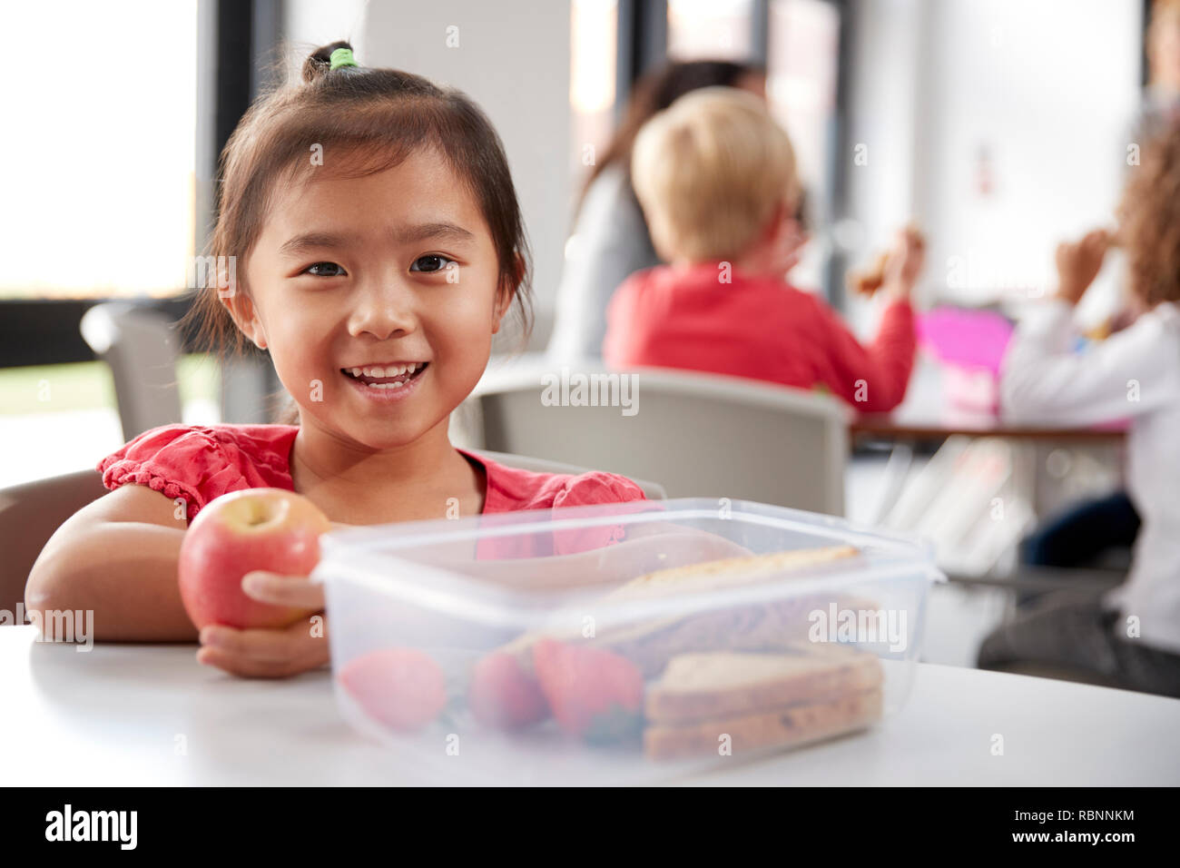 Giovani cinesi schoolgirl seduti ad un tavolo durante la sua sosta per pranzo presso il kindergarten, tenendo un apple e sorridente, close up Foto Stock