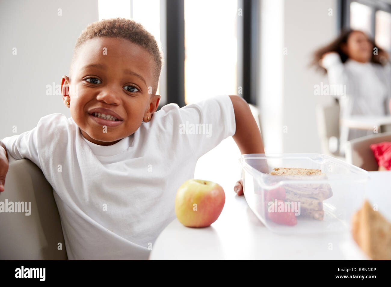 Giovane nero schoolboy seduti ad un tavolo sorridente in un asilo nido classe durante la sua pausa pranzo, close up Foto Stock