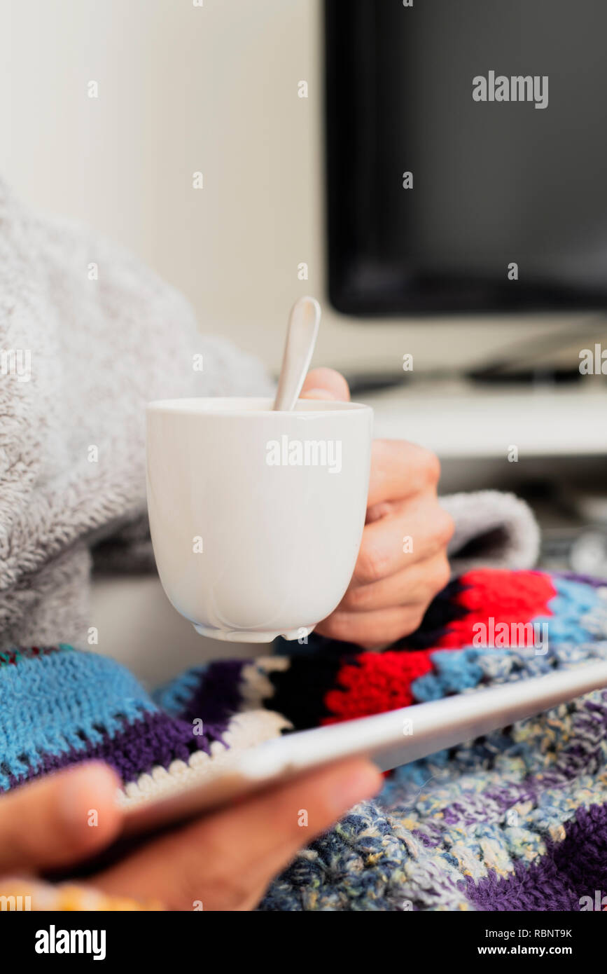 Primo piano di un malato giovane uomo caucasico a casa indossando una casa soffice accappatoio e avvolto in un colorato lavorate a maglia coperta, il riscaldamento con una tazza di zuppa calda Foto Stock