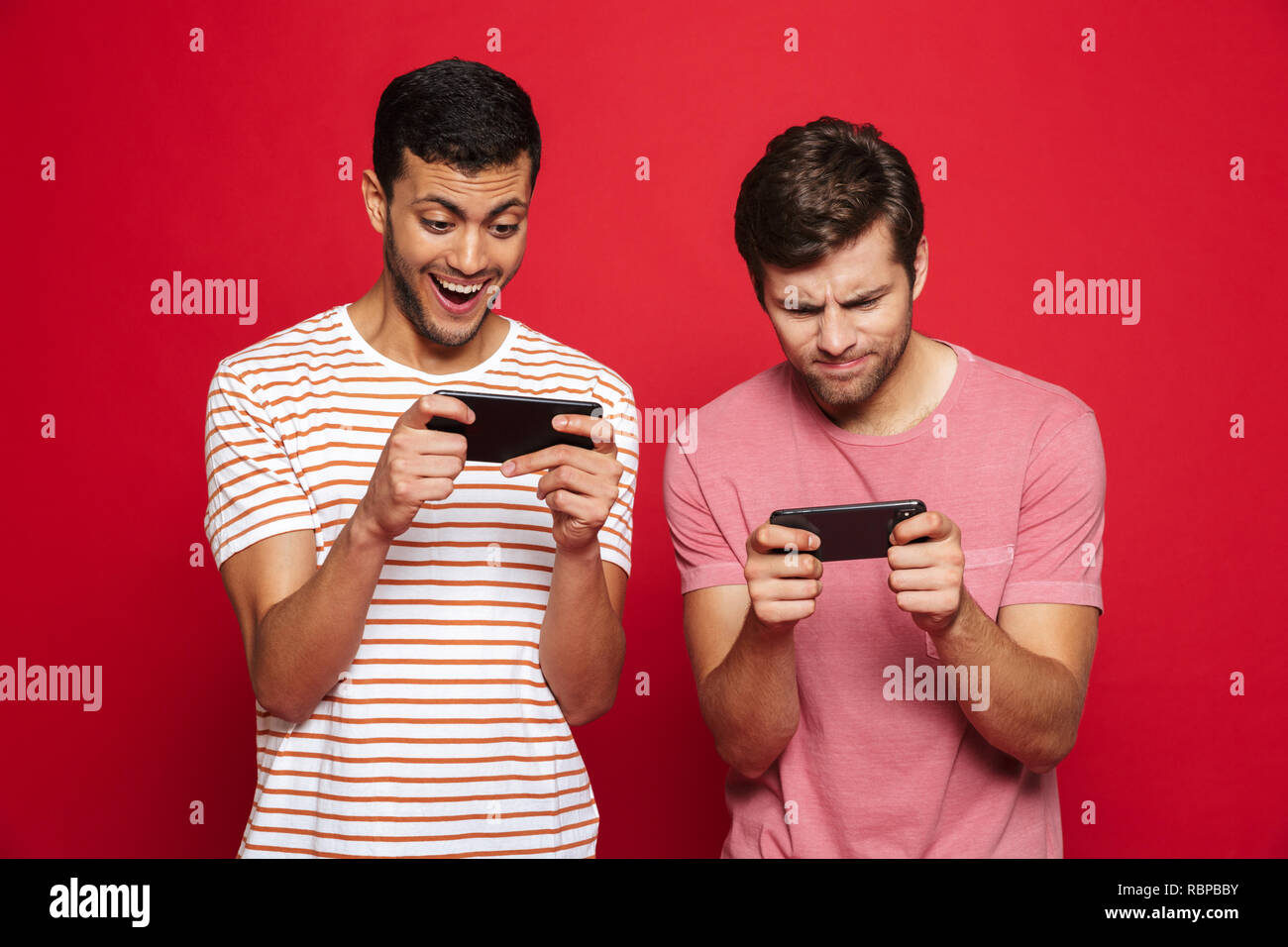 Due allegro giovani uomini in piedi isolato su sfondo rosso, giochi sul telefono cellulare Foto Stock