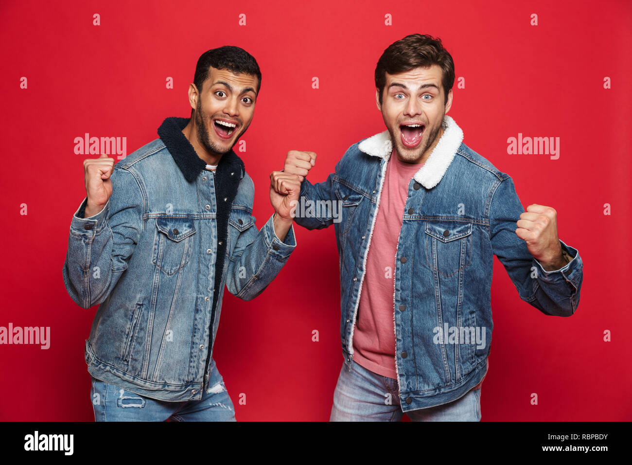 Due entusiasti giovani amici indossando giubbotti denim in piedi isolato su sfondo rosso, celebrando Foto Stock