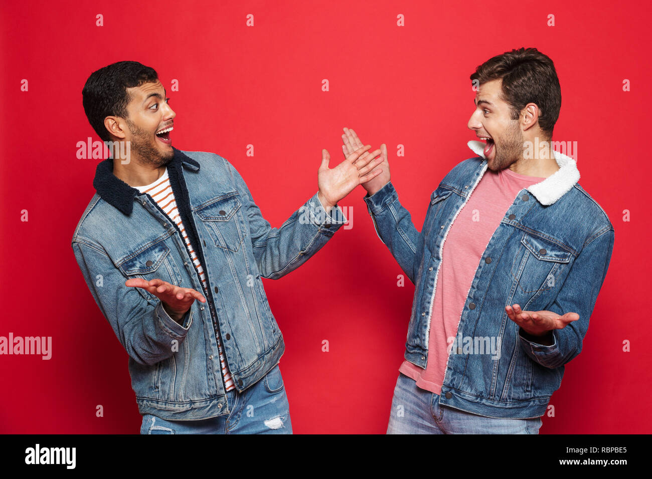 Due entusiasti giovani amici indossando giubbotti denim in piedi isolato su sfondo rosso, celebrando Foto Stock