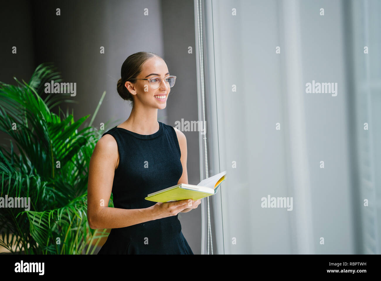 Ritratto di una giovane e bella ed elegante di donna bionda con gli occhiali per leggere un libro da una finestra durante il giorno. Ella è professionalmente vestito. Foto Stock