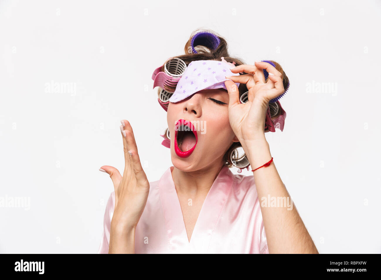 Stanco casalinga con bigodini nei capelli indossare accappatoio sbadigli isolate su sfondo bianco, indossa una maschera di sospensione Foto Stock