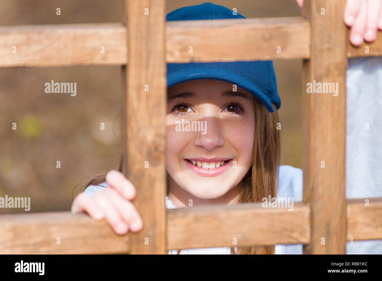 Indossando un tappo a sfera e un grande sorriso di una giovane ragazza guarda la fotocamera da behine un quadrato in legno a traliccio. Foto Stock