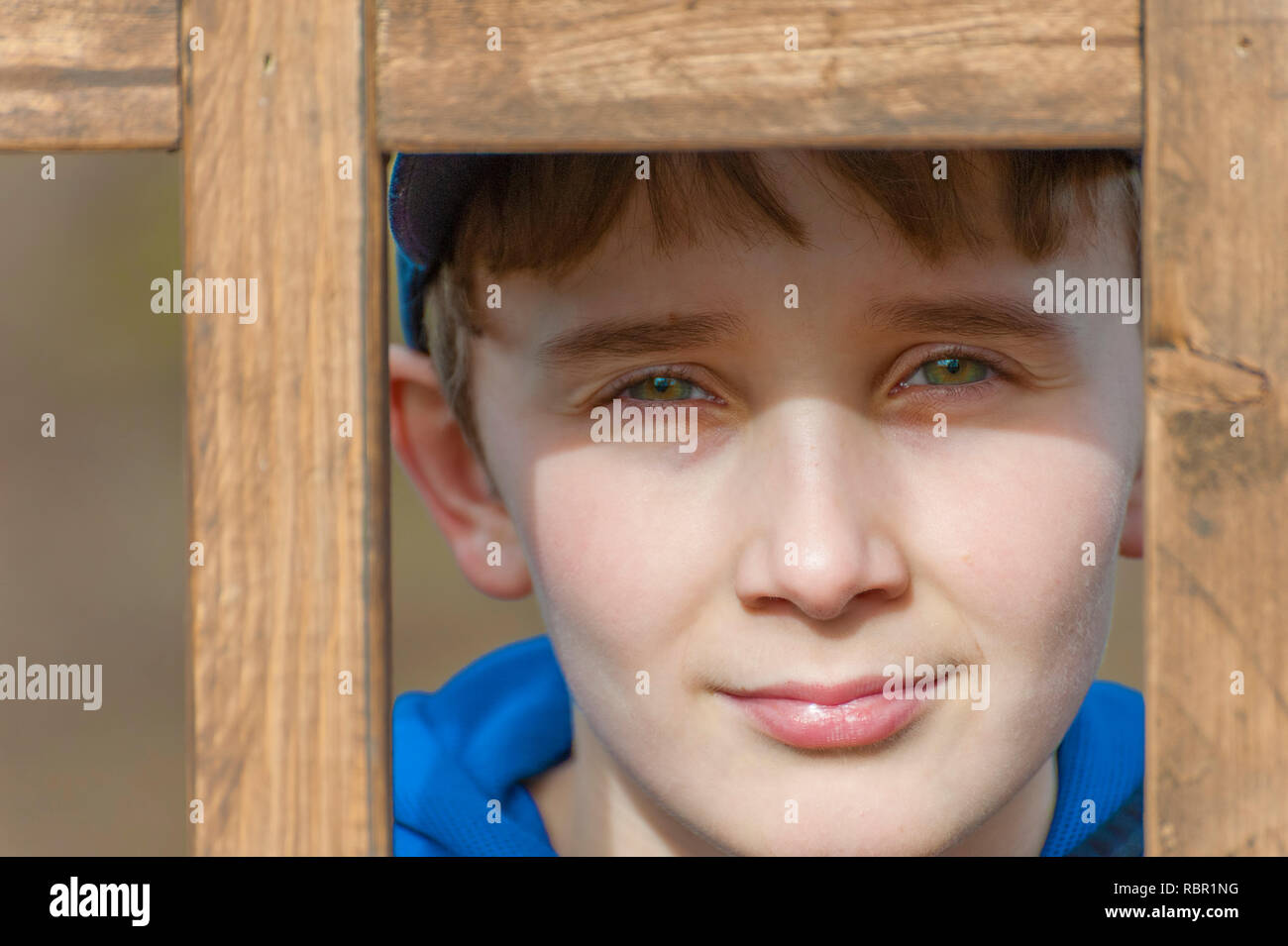 Un giovane ragazzo con bellissimi occhi verdi guarda la fotocamera da dietro un latice in legno. Foto Stock