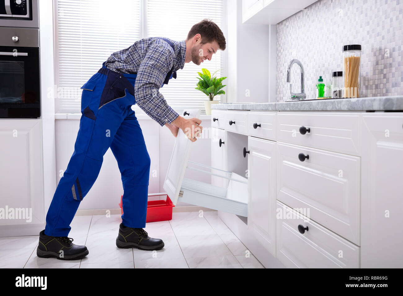 Vista laterale di un giovane tuttofare In uniforme di installare il cassetto in cucina Foto Stock