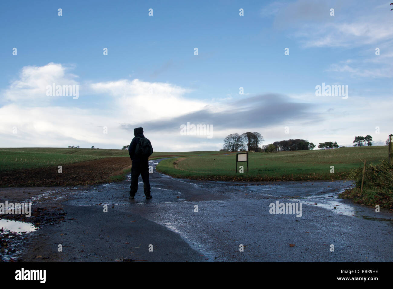 Un solitario escursionista incappucciato con uno zaino di fronte ad un bivio su un inverni giornata in campagna. Foto Stock