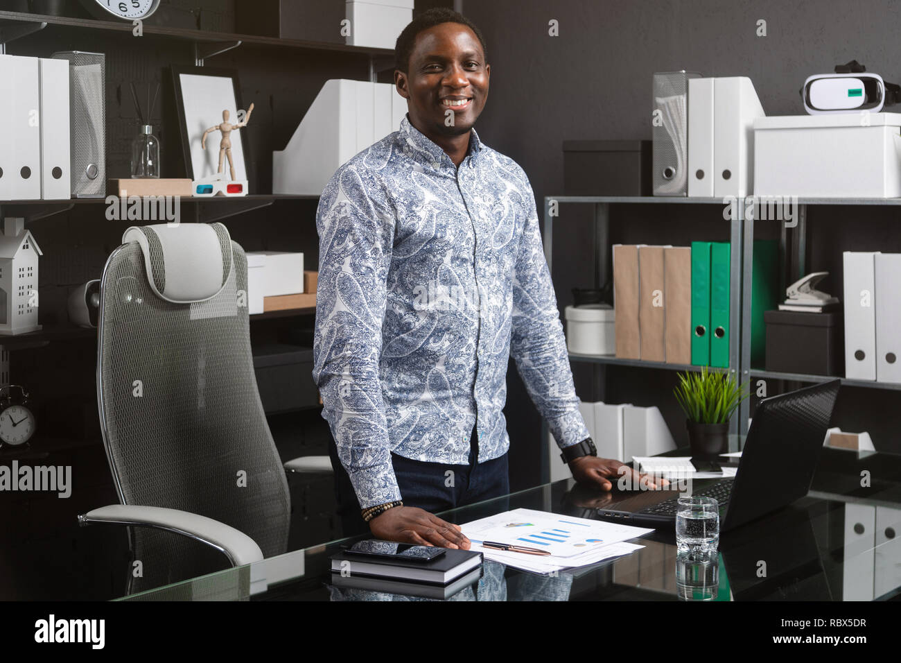 Ritratto di tre quarti lungo giovane imprenditore nero in piedi vicino alla scrivania in ufficio Foto Stock