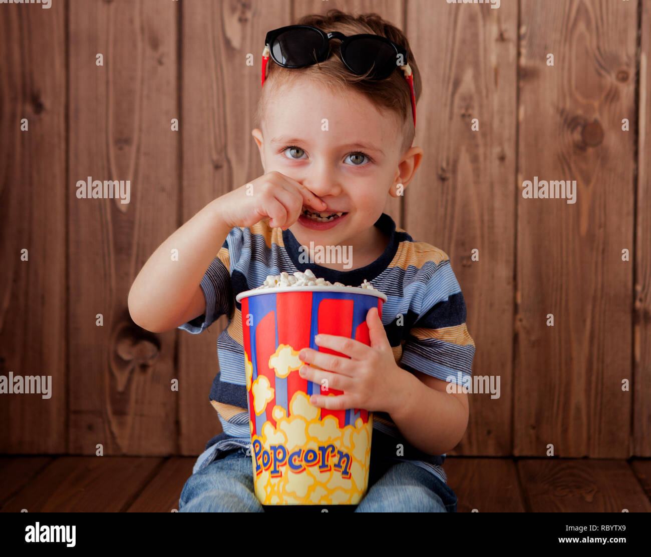 Poco carino kid bimbo da 2 a 3 anni , 3d cinema Imax bicchieri tenendo la benna per popcorn, mangiare fast food su sfondo di legno. I bambini la vita dell infanzia Foto Stock