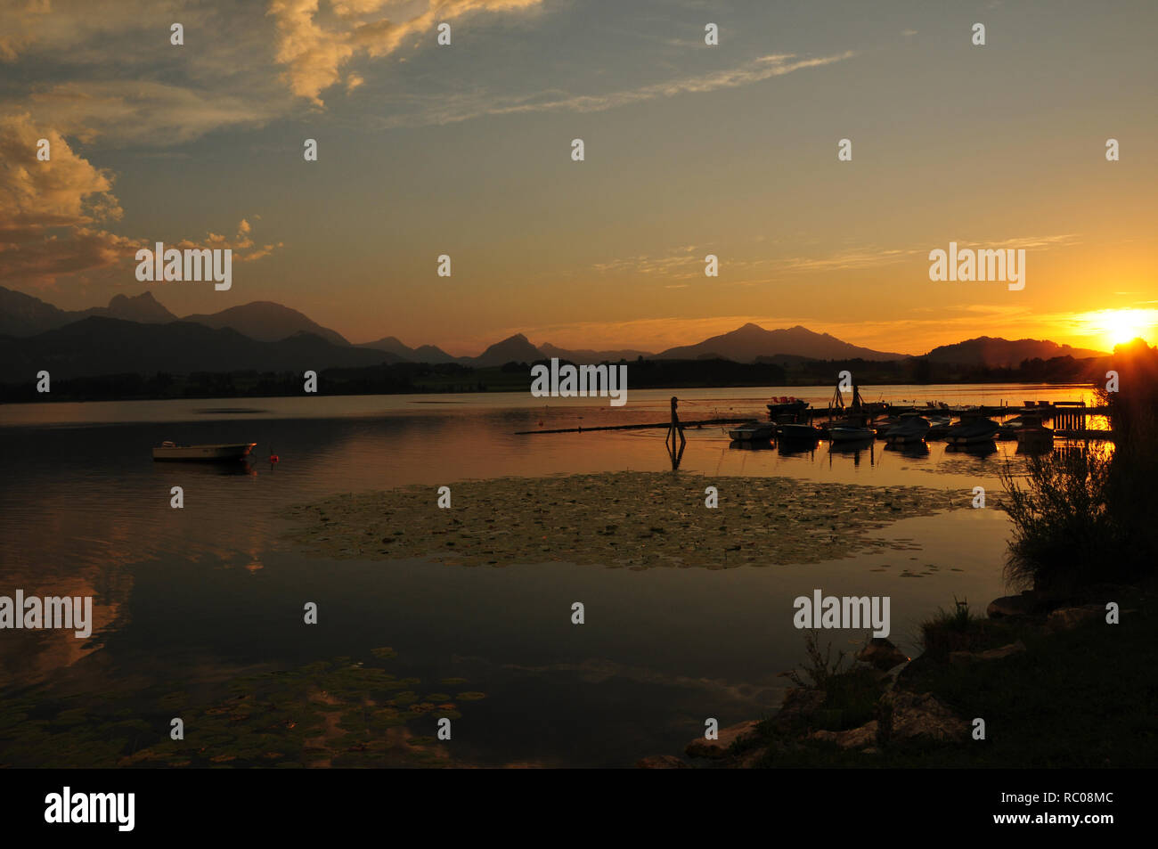 La superficie riflettente del Lago di Hopfen con tre barche presso la passerella nel sole di setting Foto Stock