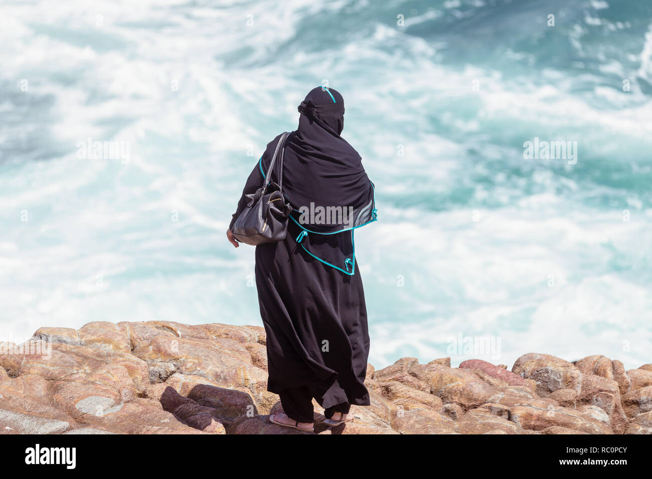 Arabo donna in abiti tradizionali guardando grandi onde in Hermanus, Sud Africa Foto Stock