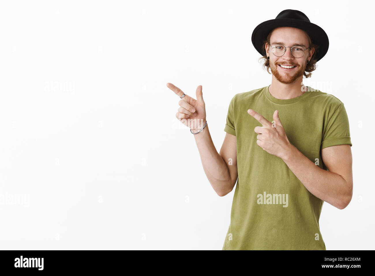 Gioioso e cordiale soddisfatto barbuto hipster uomo nel cappello e occhiali con naso forato rivolto ad angolo superiore sinistro con il dito pistole e sorridente largamente in telecamera su uno sfondo grigio Foto Stock