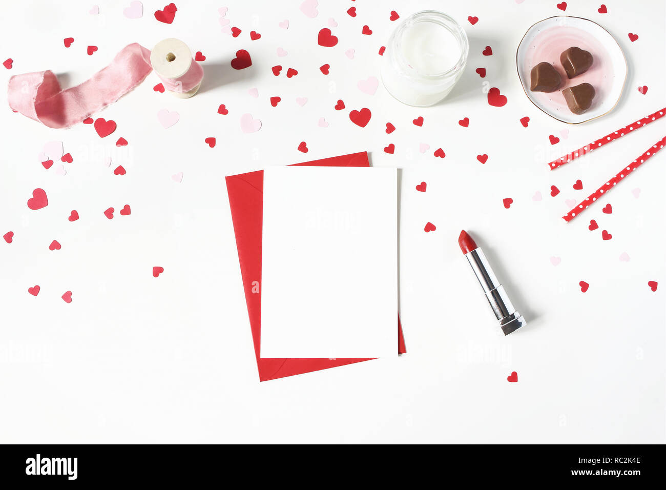 Il giorno di San Valentino, wedding still life scena. Candela, carta coriandoli, Cioccolato cuori, rossetto e bigliettino vuoto mockup sul tavolo bianco sullo sfondo. Il concetto di amore. Appartamento laico, vista dall'alto. Foto Stock