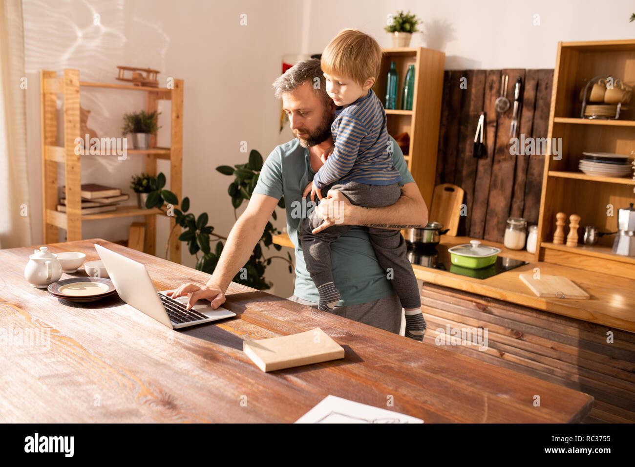 Considerato grave bello barbuto unico padre holding piccolo figlio e digitando su laptop mentre si lavora a casa Foto Stock