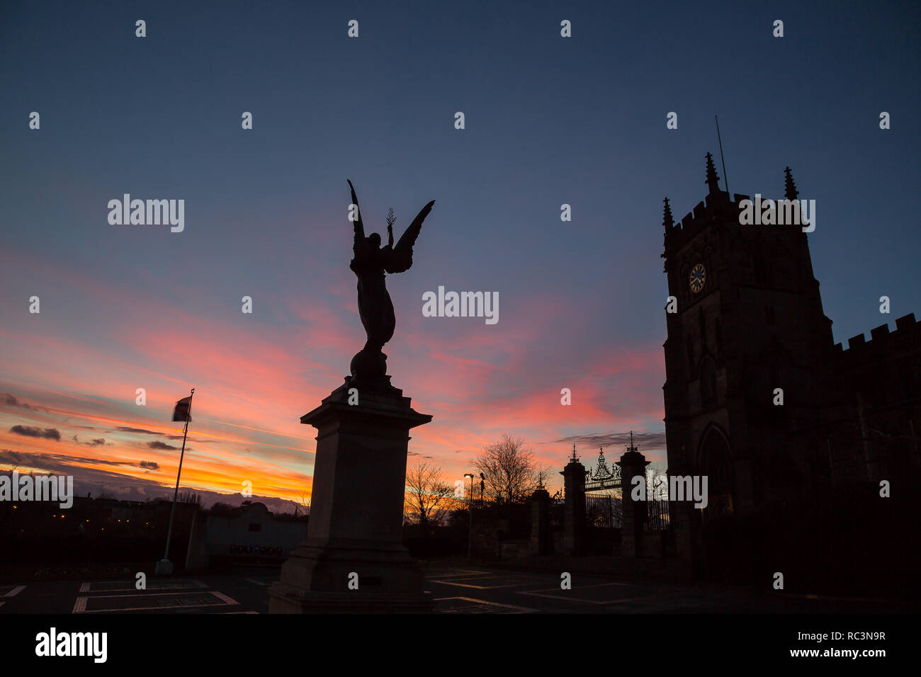 Kidderminster, Regno Unito. Il 13 gennaio, 2019. Regno Unito: meteo il sole tramonta a Kidderminster dopo un insolitamente calda giornata con temperature che raggiungono i 10 gradi celsius. La scultura di un angelo di pace può essere visto qui retroilluminati da il tramonto colorato al di fuori di St Mary's Church, Kidderminster. Credito: Lee Hudson/Alamy Live News Foto Stock