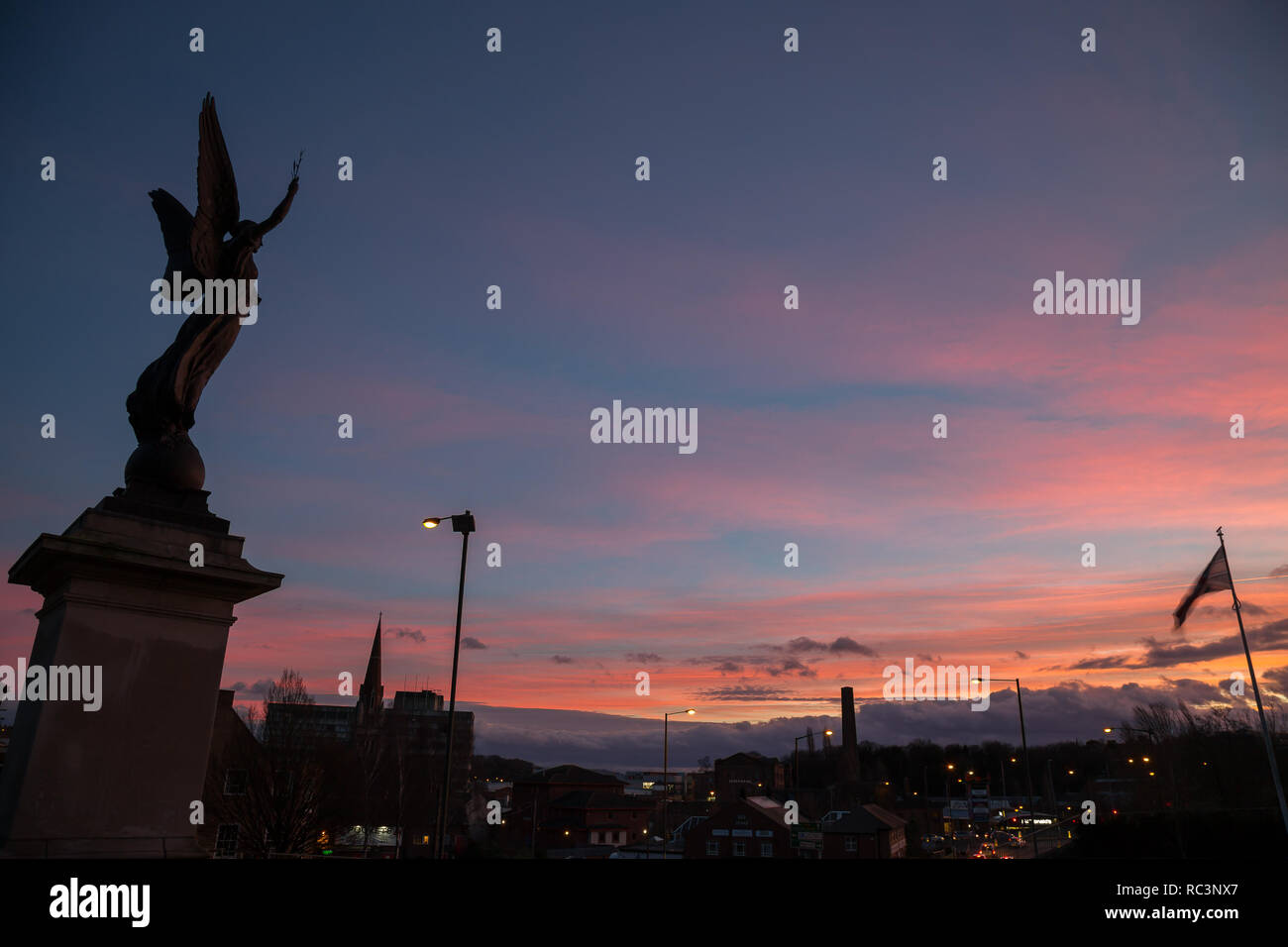 Kidderminster, Regno Unito. Il 13 gennaio, 2019. Regno Unito: meteo il sole tramonta a Kidderminster dopo un insolitamente calda giornata con temperature che raggiungono i 10 gradi celsius. La scultura di un angelo di pace può essere visto qui retroilluminati da il tramonto colorato al di fuori di St Mary's Church, Kidderminster. Credito: Lee Hudson/Alamy Live News Foto Stock