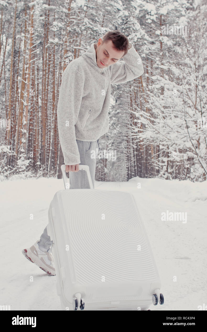 Ritratto di giovane uomo alla moda in bianche vesti, passeggiate sulla neve tenendo la valigia e andare via pineta. Vacanza invernale concetto di viaggio. Outdoo Foto Stock