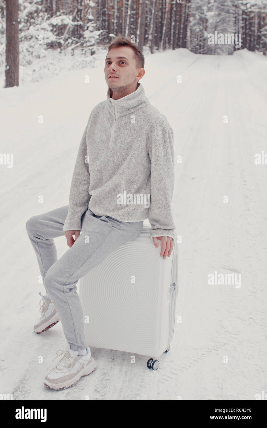 Ritratto di giovane uomo alla moda in bianche vesti, passeggiate sulla neve tenendo la valigia e andare via pineta. Vacanza invernale concetto di viaggio. Outdoo Foto Stock