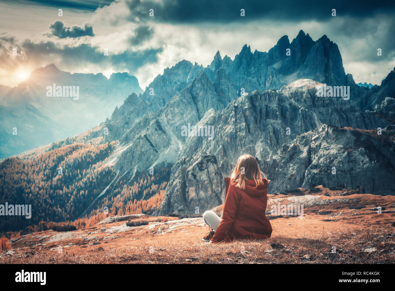 Giovane donna è seduta sulla collina contro le maestose montagne al tramonto in autunno nelle Dolomiti, Italia. Paesaggio con ragazza, cielo nuvoloso, orange gra Foto Stock