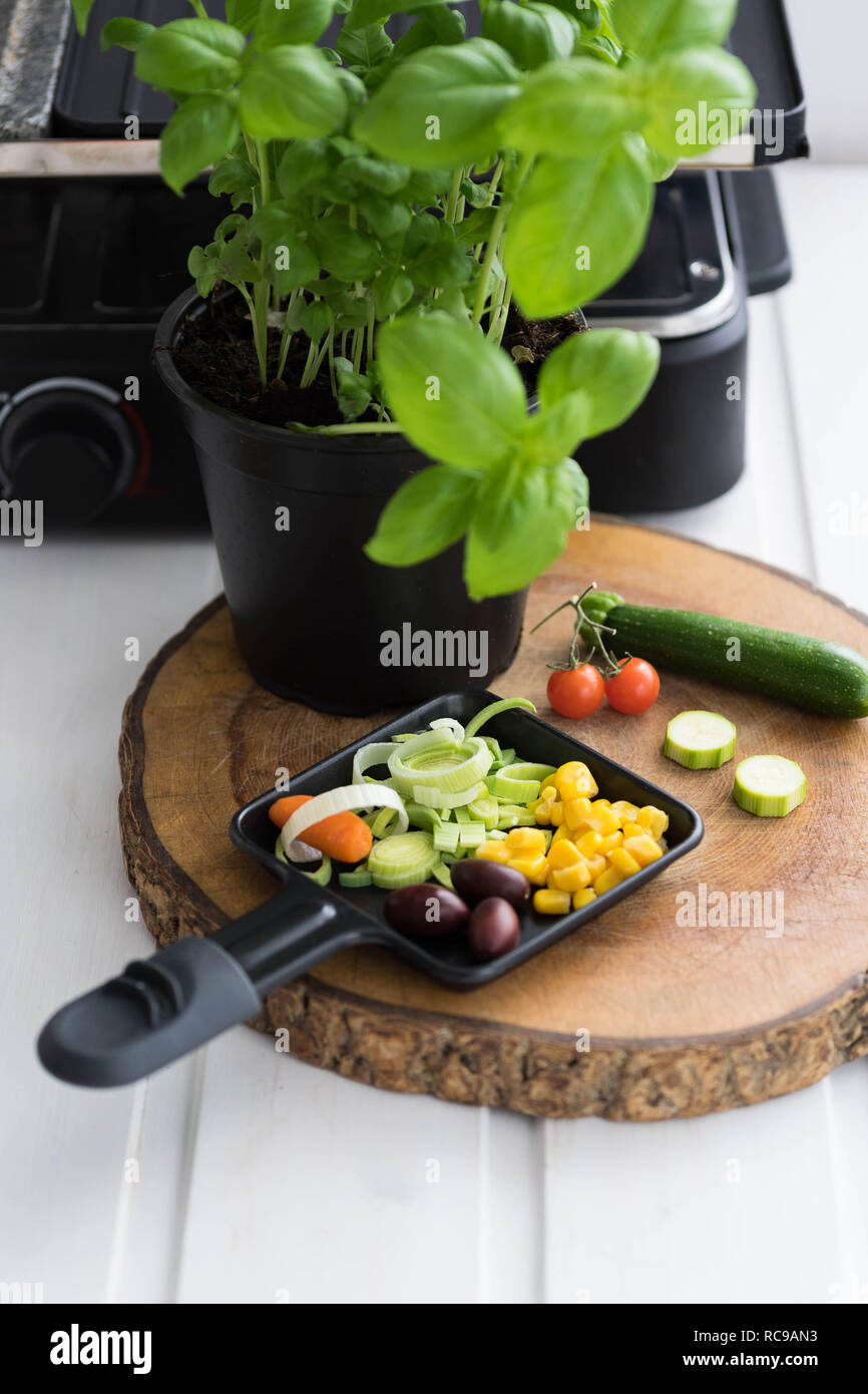 Vegan raclette, raclette pan con un trito di verdure su una tavola di legno accanto alle zucchine e una pentola di basilico Foto Stock