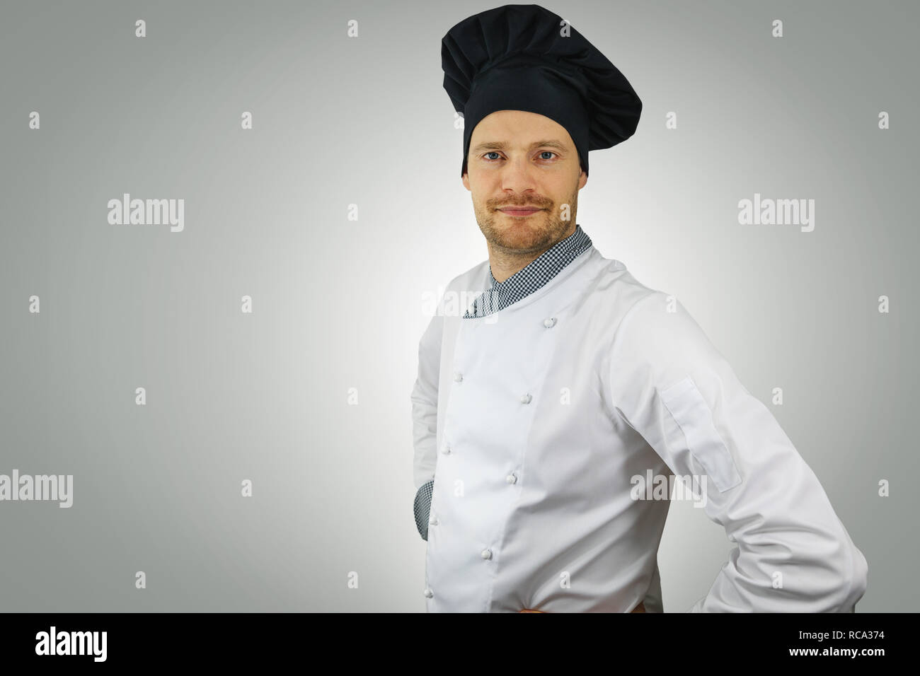 Il giovane chef in uniforme con le mani sui fianchi in piedi su sfondo grigio Foto Stock