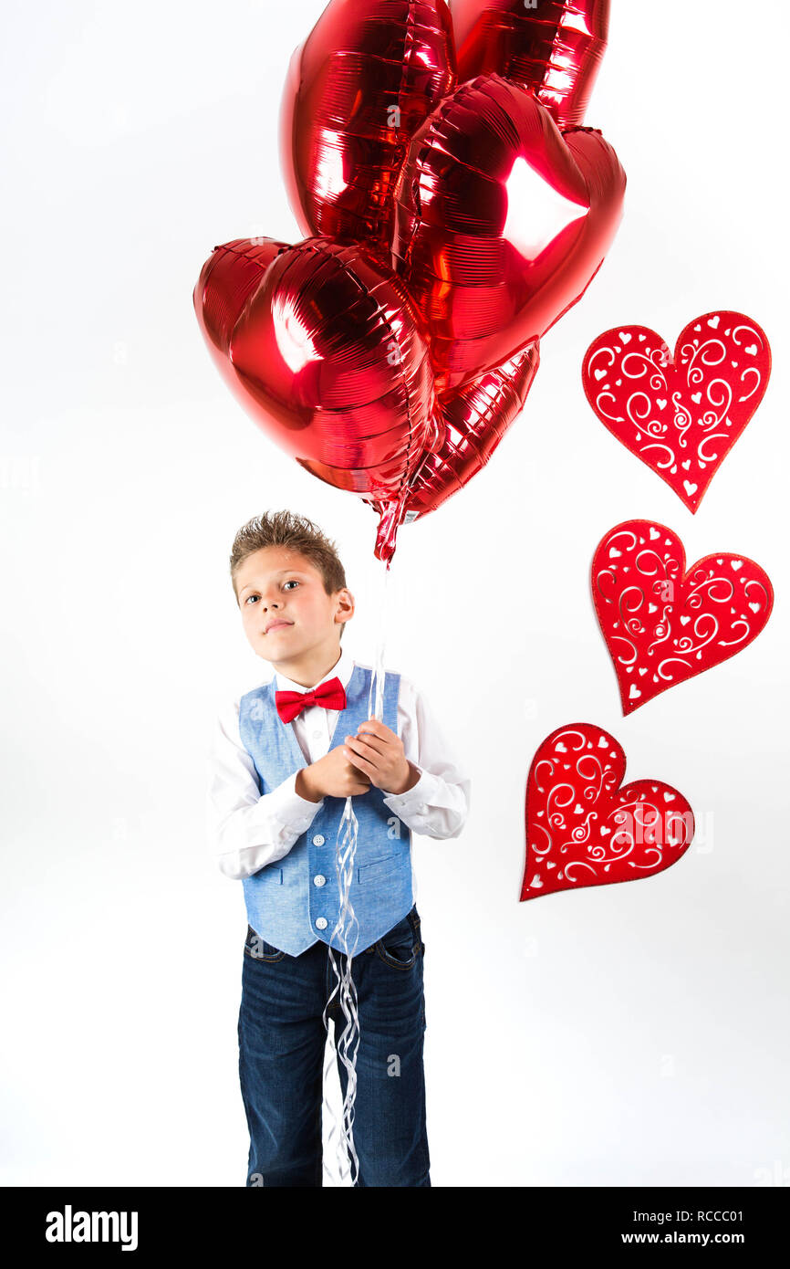 Bello piccolo ragazzo con abito bianco per celebrare il giorno di San Valentino e azienda lucido rosso palloncini a forma di cuore su sfondo bianco Foto Stock