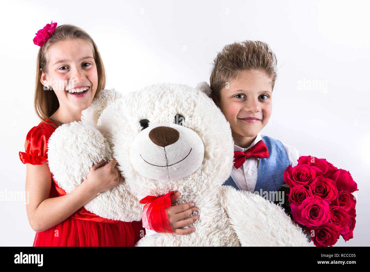 Grazioso e piccolo ragazzo e una ragazza che celebra il giorno di San Valentino e azienda rose rosse benna e orso bianco dono sullo sfondo bianco con cuore rosso Foto Stock