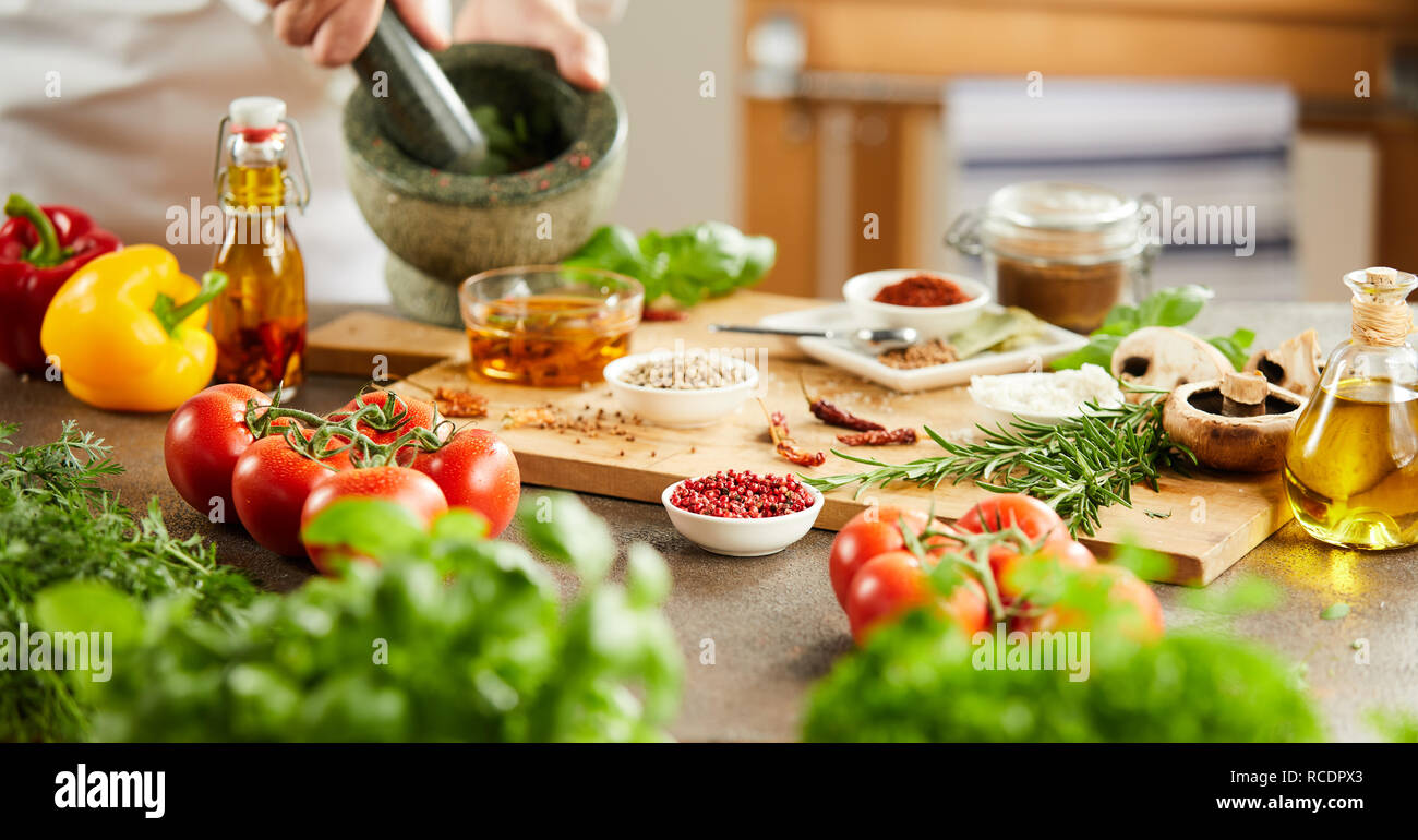 Banner panorama delle mani di uno chef che prepara le erbe miscelando in un pestello e mortaio con assortimento di spezie e verdure fresche su una tavola di legno b Foto Stock