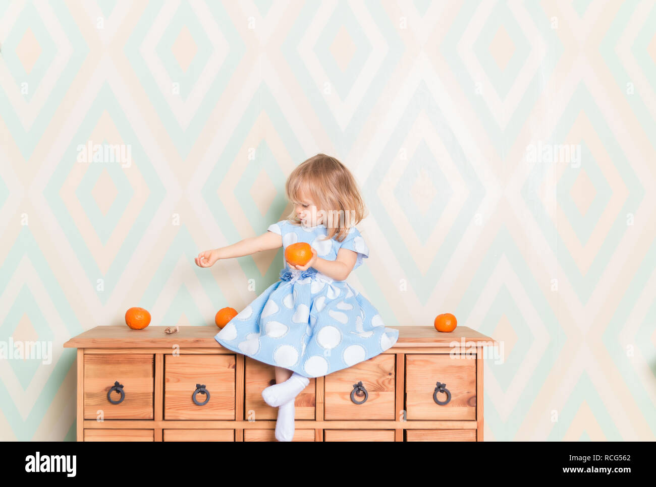 Ritratto di blonde Caucasian bambina con gli occhi blu in abito elegante e calzini bianchi seduta sul comò in legno con mandarino clementina frutto in mano e guardando dal lato a mano. Lo stupore Foto Stock