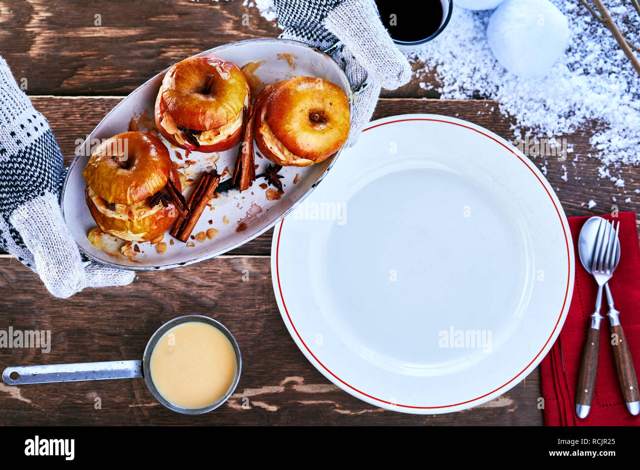 Mani guantate tenendo un piatto di sano dessert invernale di insaccati mele cotte con cannella spice su una tabella innevato con la piastra a vuoto e crema pasticcera sau Foto Stock