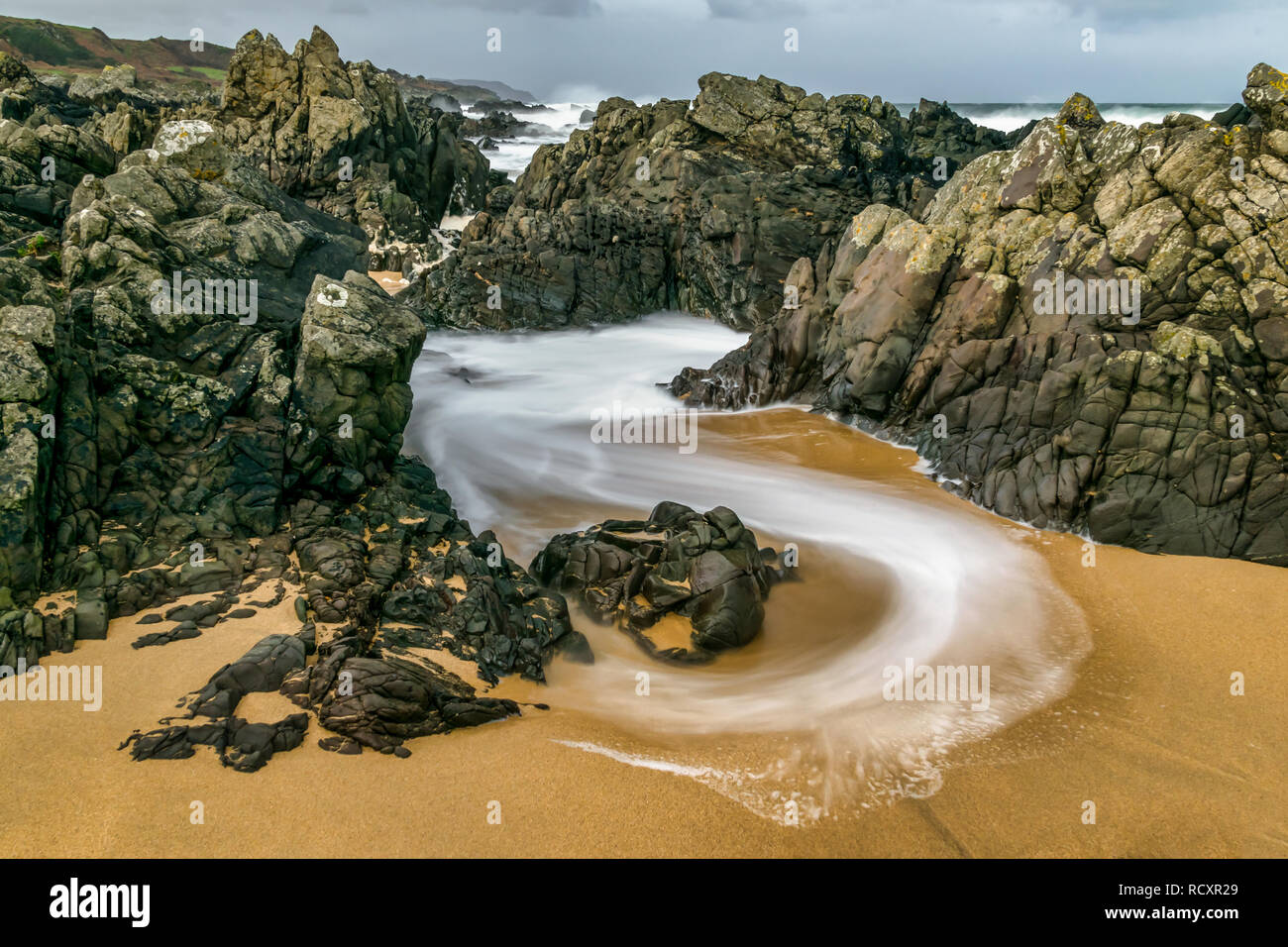 Bassa velocità dell'otturatore è stata utilizzata per catturare questa immagine di un vorticoso d'onda se le rocce sulla spiaggia. L'acqua sfuggente creato curve di schiuma bianca Foto Stock