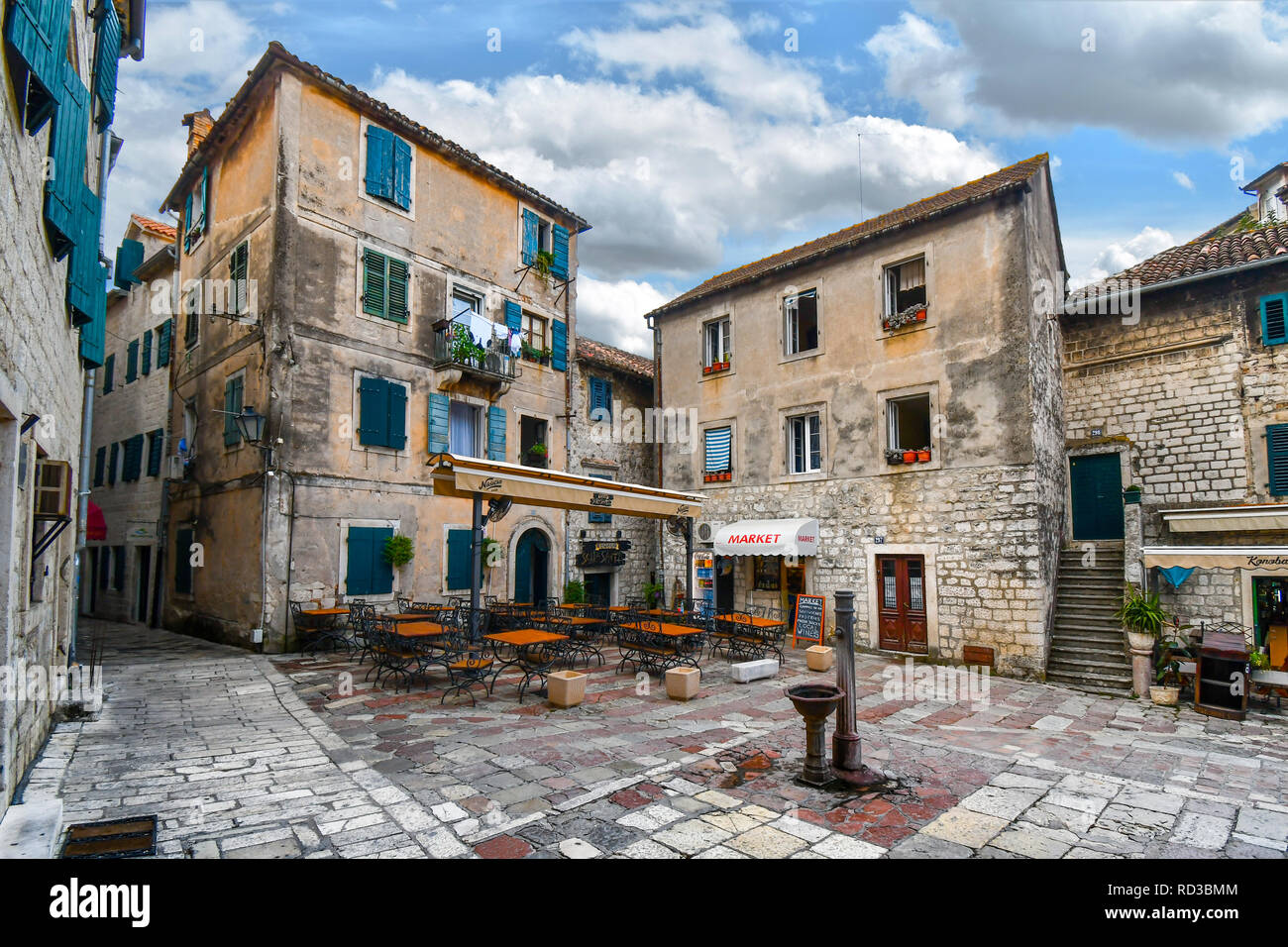 Una piccola piazza nella città vecchia di Kotor, Montenegro, con una pompa ad acqua nel centro circondato da un cafe, mercato e appartamenti con vetri colorati. Foto Stock