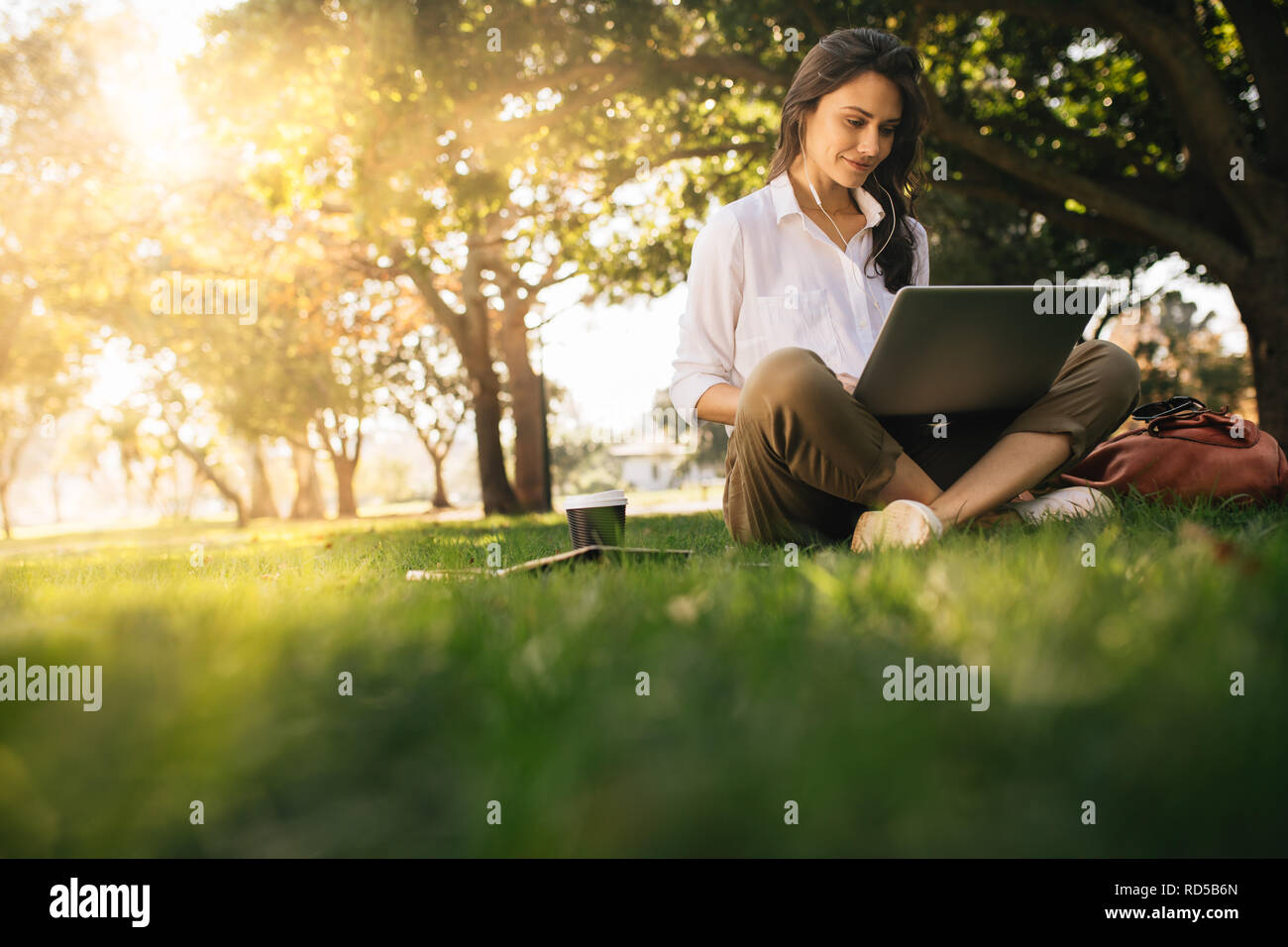 Donna seduta su erba a park lavorando sul computer portatile. Donna che indossa gli auricolari utilizzando computer portatile mentre seduto sotto un albero a parco con condizioni di luce solare intensa da b Foto Stock