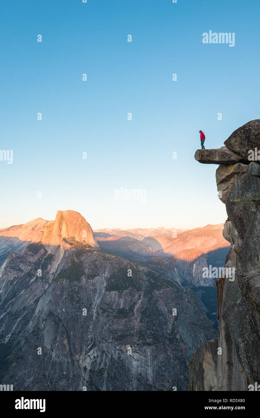 Un intrepido escursionista è in piedi su una roccia a strapiombo godendo della vista verso il famoso Half Dome presso il Glacier Point si affacciano nella bellissima crepuscolo serale Foto Stock