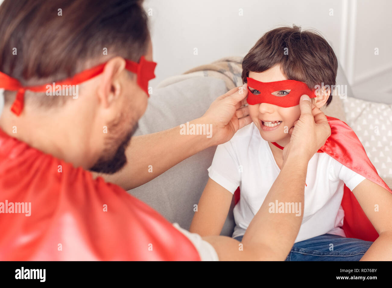 Padre e figlio in costumi superheroe a casa seduti sul divano uomo maschera mettendo sul ragazzo che sorride close-up Foto Stock