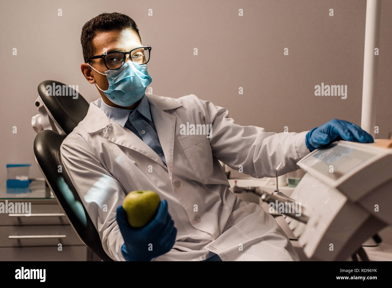 African American dentista seduto in bicchieri e maschera e azienda apple Foto Stock