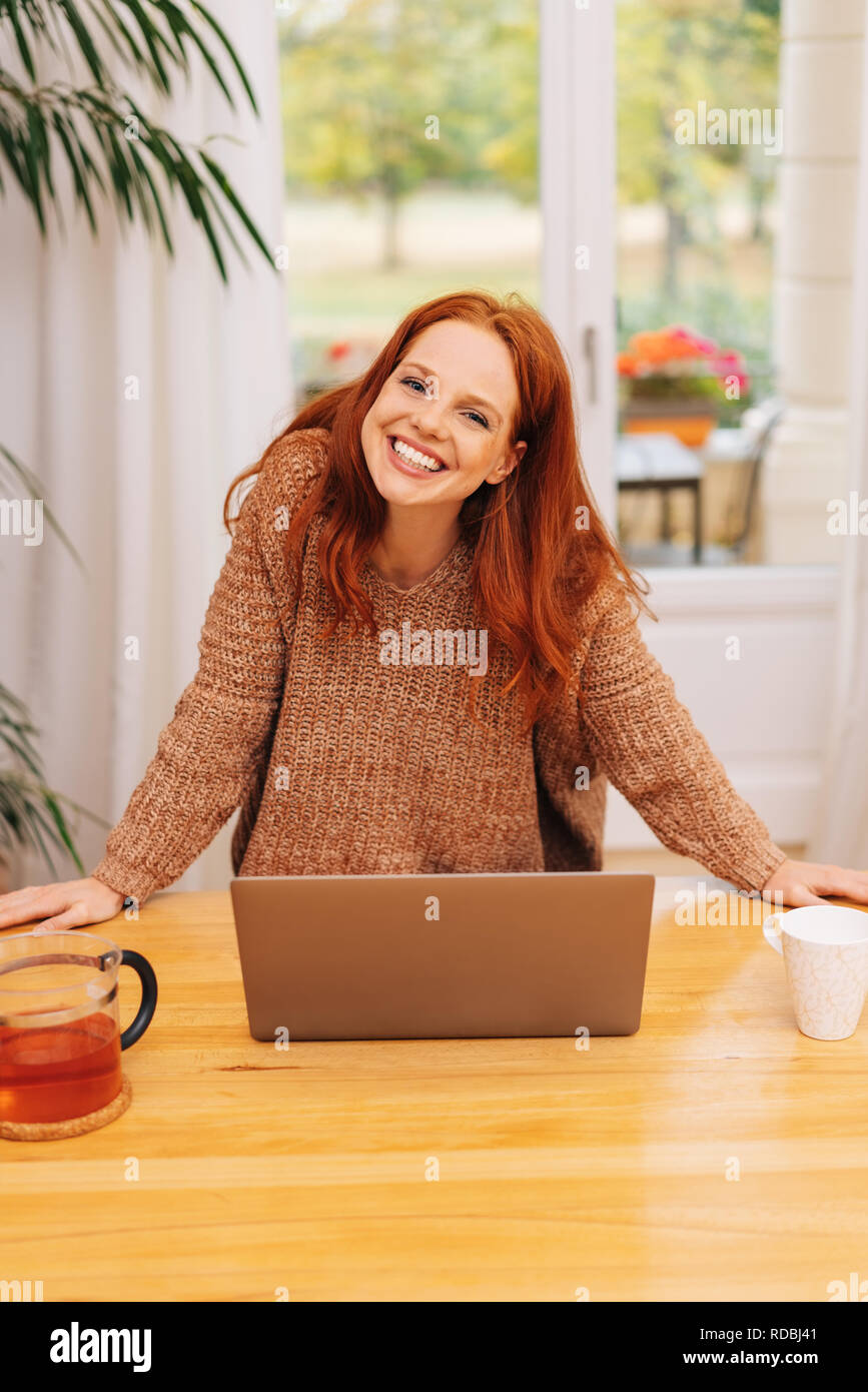 Gentile sorridente giovane i capelli rossi donna in felpa lavorata a maglia in piedi e appoggiato sul tavolo con laptop e guardando la fotocamera. Parte anteriore a metà lunghezza ritratto Foto Stock