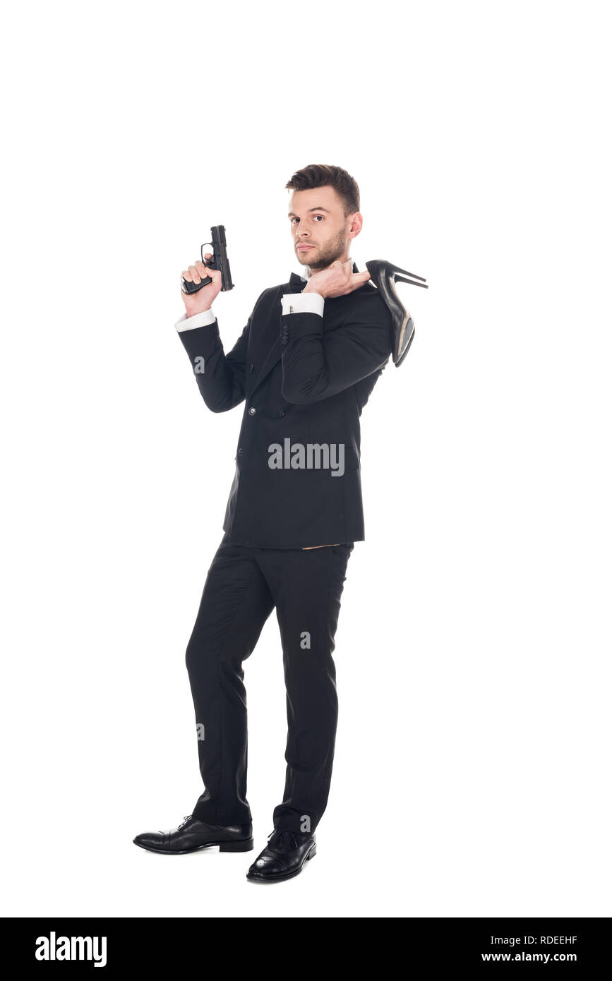 Elegante agente segreto in abito nero azienda pistola e tacchi alti, isolato su bianco Foto Stock