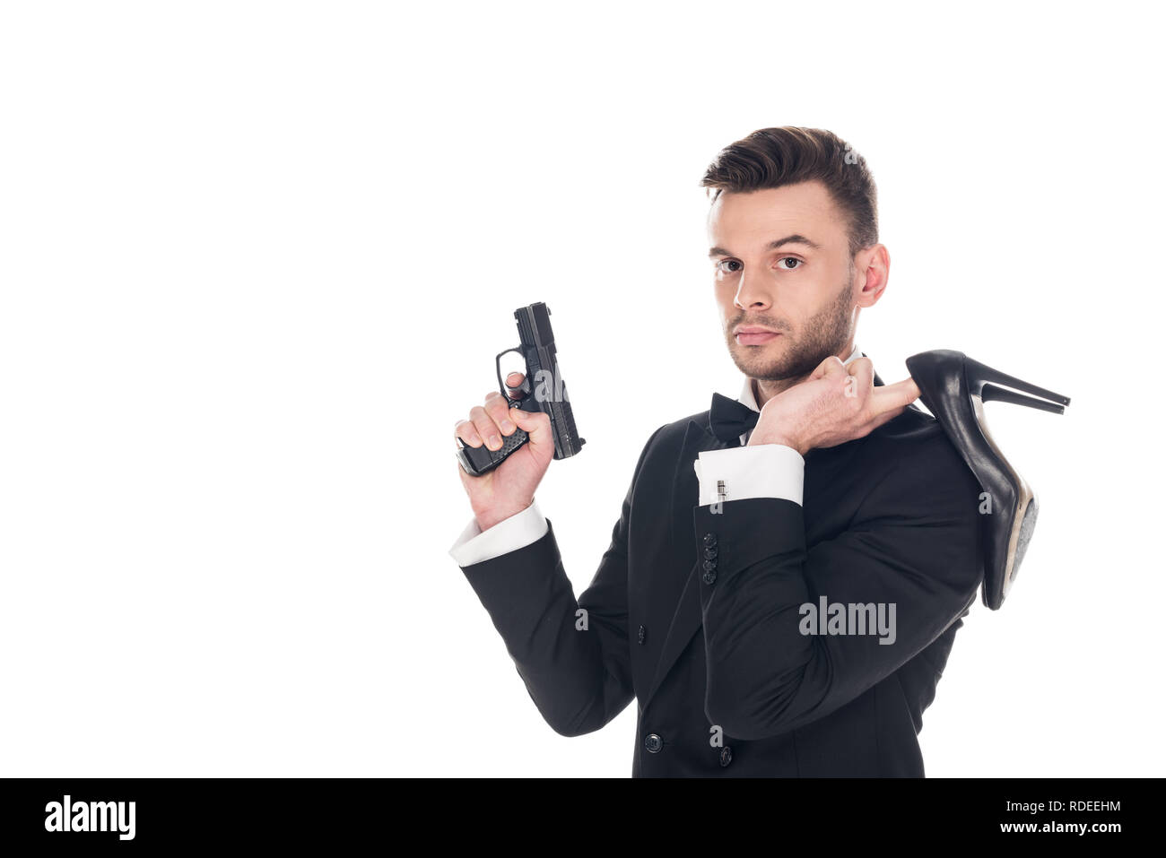 Bello agente segreto in abito nero azienda pistola e tacchi alti, isolato su bianco Foto Stock