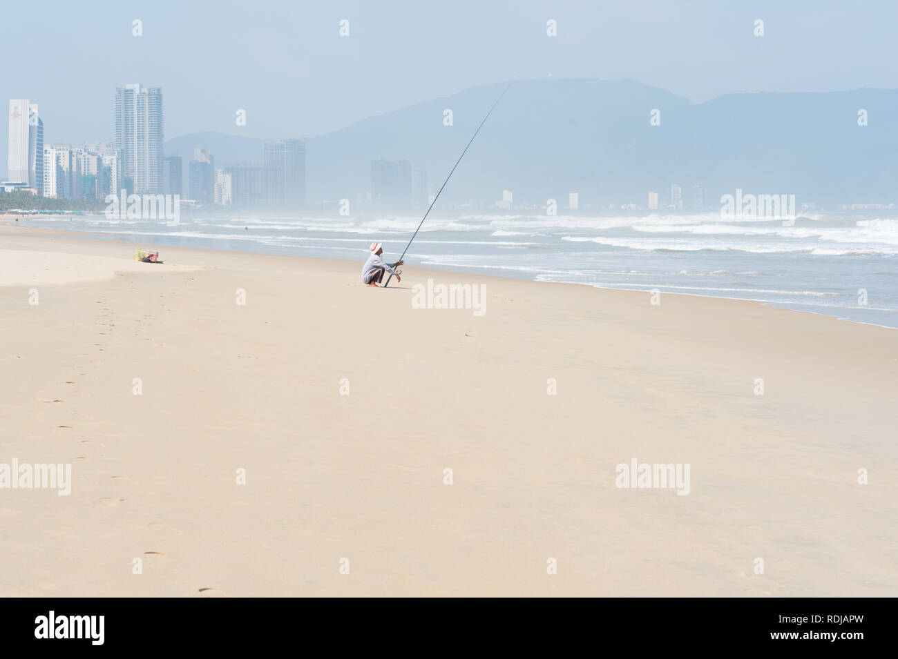 Danang, Vietnam - 1 Novembre 2018: un uomo pesci con un asta di filatura nel profondo mare ondeggiante seduto sulla soleggiata spiaggia vuota con Danang city dietro Foto Stock