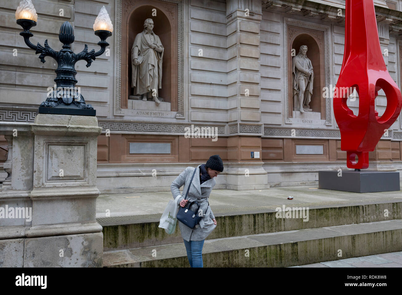 Una donna controlla i suoi vestiti dopo la seduta sotto la statua di British filosofo Vittoriano John Locke (da William Theed) e una scultura moderna di Renzo Piano, con un full-size riproduzione in fibra di vetro di un 'gerberette', una delle die-cast rocker travi a sbalzo da Parigi' Georges Pompidou edificio, il 10 gennaio 2019, a Londra, in Inghilterra. Il filosofo inglese John Locke (1632 - 1704) il cui effigi sono da William Theed, noto anche come William Theed, il giovane (1804 - 9 settembre 1891), scultore inglese i cui servizi sono stati ampiamente utilizzati dalla famiglia reale. Foto Stock