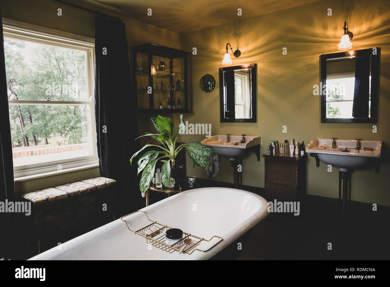 Vista interna del bagno con specchi neri su vittoriano due lavandini, finestra ad anta e roll top bagno con vasca da bagno in ottone caddy. Foto Stock