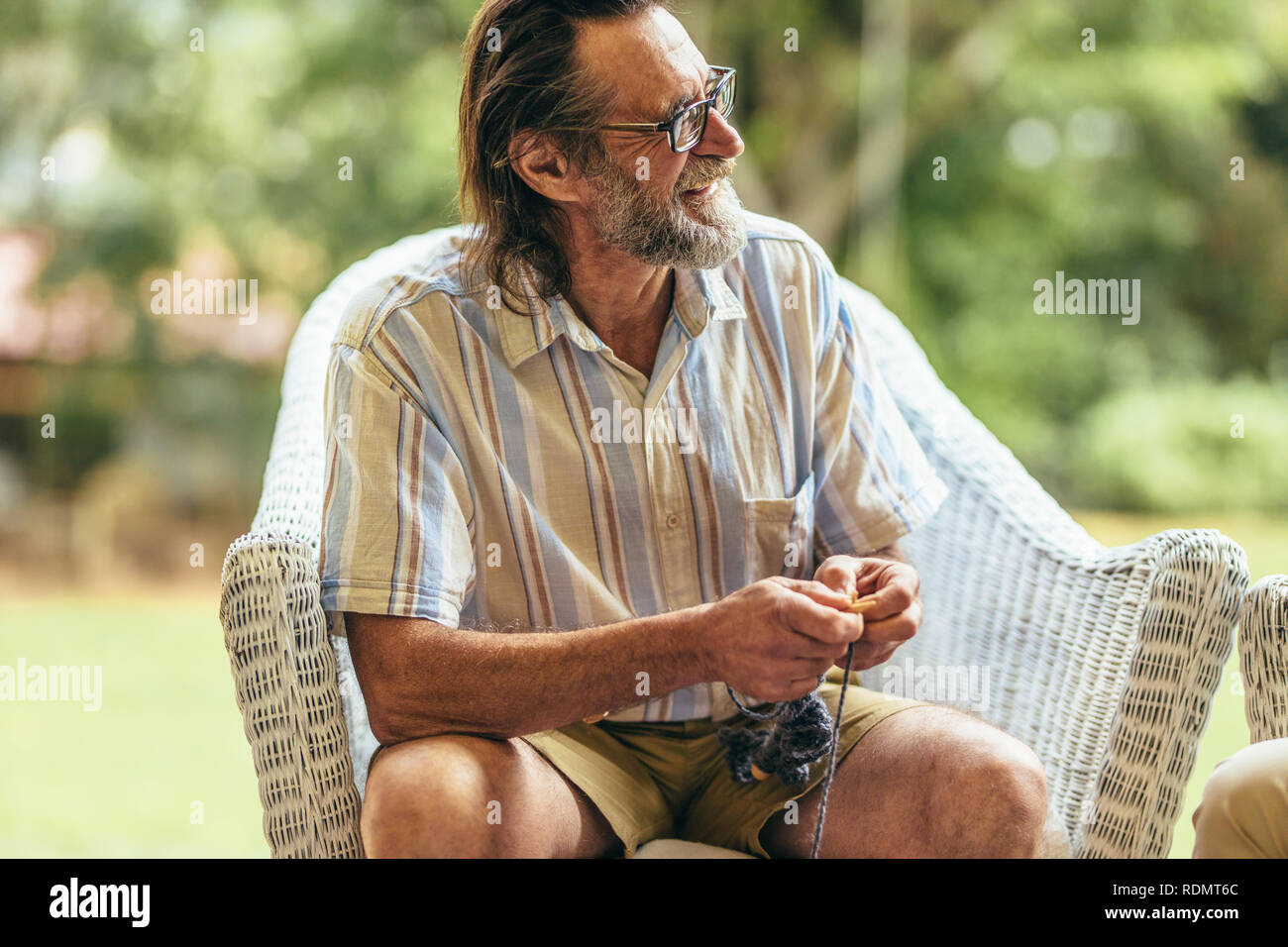 Sorridente uomo vecchio con la barba seduto su una sedia con ago da maglieria e filati di lana. Uomo in pensione rilassante sulla poltrona guardando lontano e sorridente. Foto Stock