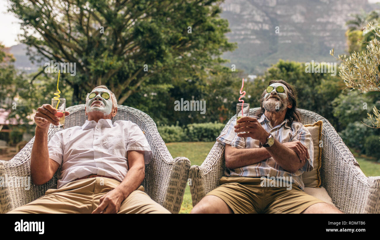 Uomini anziani con maschera al viso e fette di cetriolo sul loro volto a spa. Amici pensionati godendo del viso trattamento termale con bicchieri di succo di frutta in mano. Foto Stock
