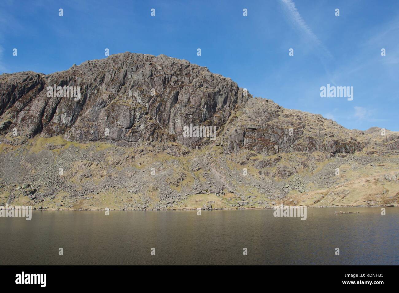 Rude scogliera di montagna si riflette ancora in acqua di un lago di montagna (tarn). Il cielo blu e drammatica pendii rocciosi. Pavey Ark, affacciato Stickle Foto Stock