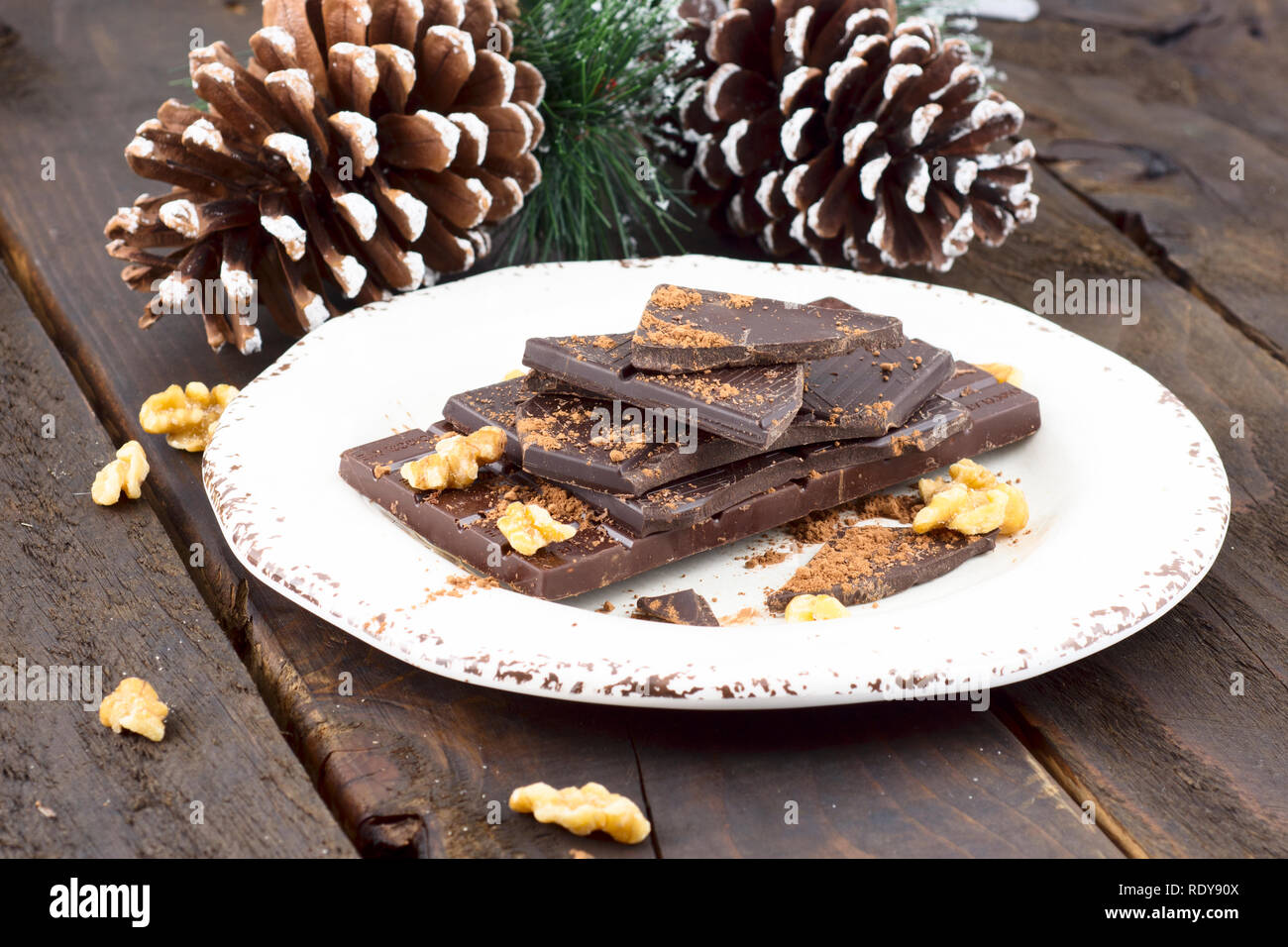 Pezzi di cioccolato e noci, su una piastra bianca, pronti per essere utilizzati per la cottura. Tavolo in legno, pigne nelle quali in background. Foto Stock