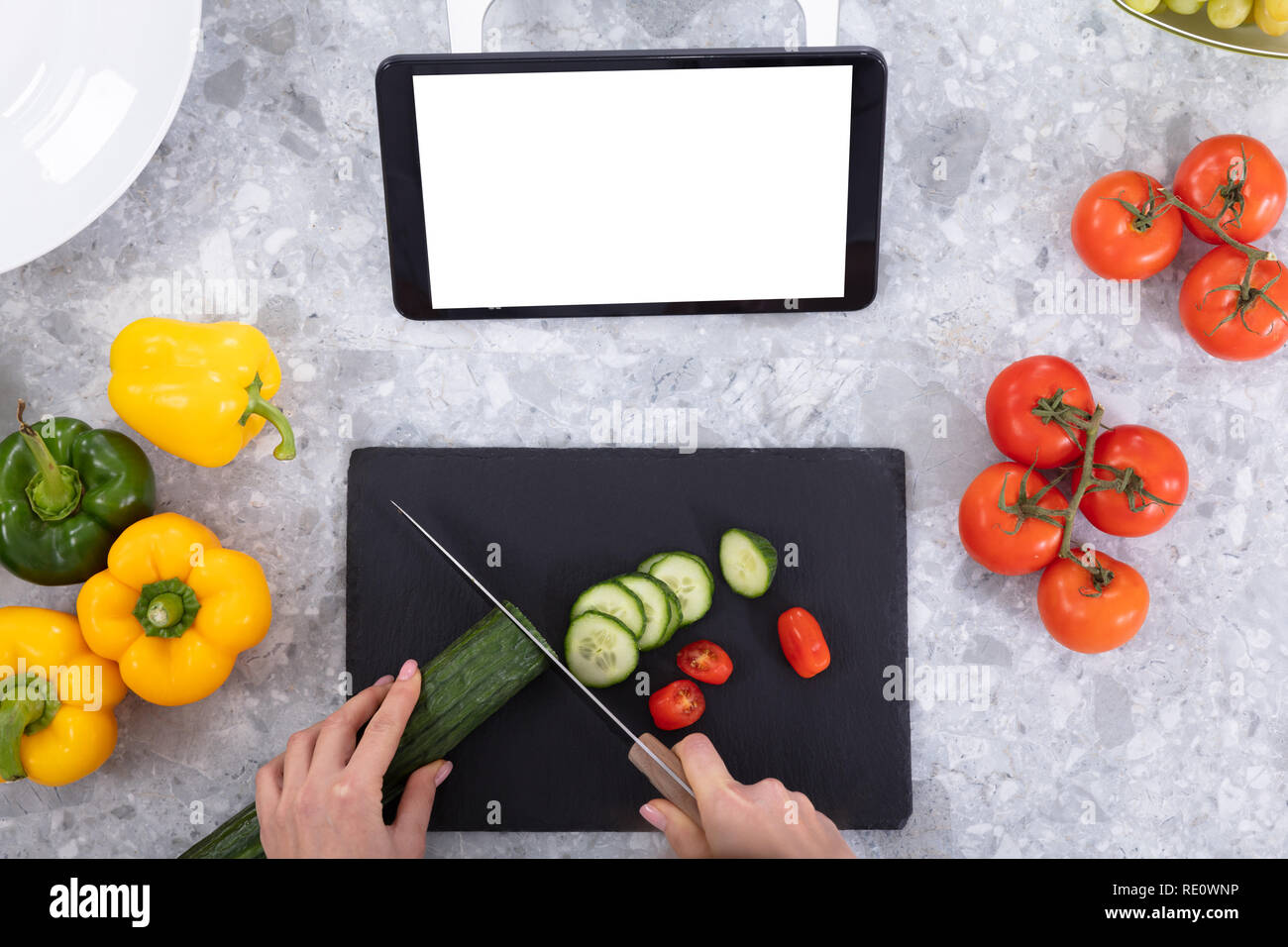 Donna cetriolo di taglio con il coltello da cucina vicino a tavoletta digitale, i pomodori e i peperoni sul banco di cucina Foto Stock